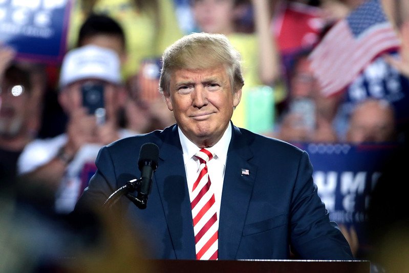 Donald Trump smiles while addressing a crowd at a political rally, showcasing campaign signs and American flags in the background.