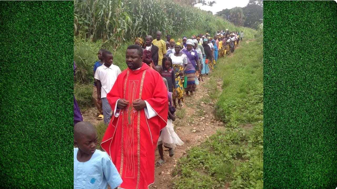 A priest in red vestments leads a procession of community members along a rural path surrounded by greenery.
