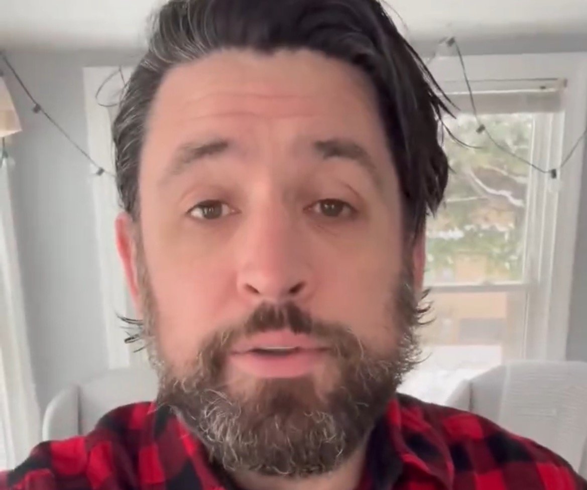 Man with a beard wearing a red and black flannel shirt speaking indoors, with snow visible outside the window.