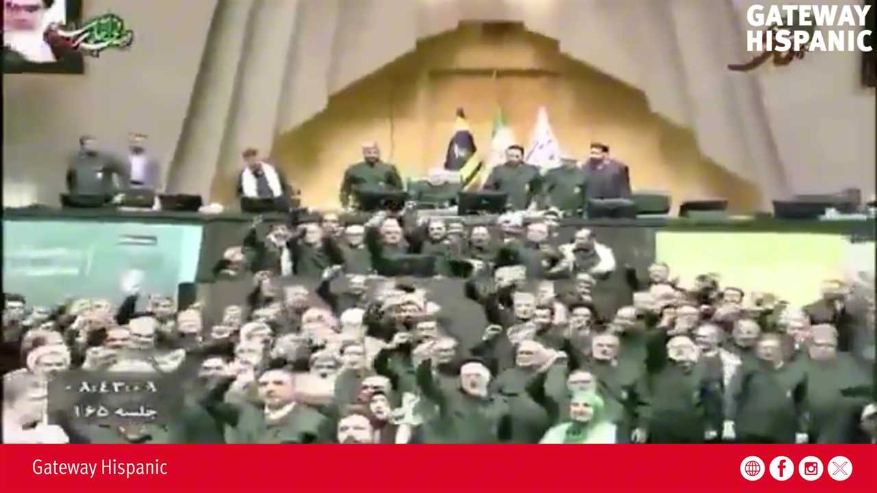 A large group of officials in dark uniforms cheer enthusiastically during a session in a legislative assembly, with flags displayed in the background.