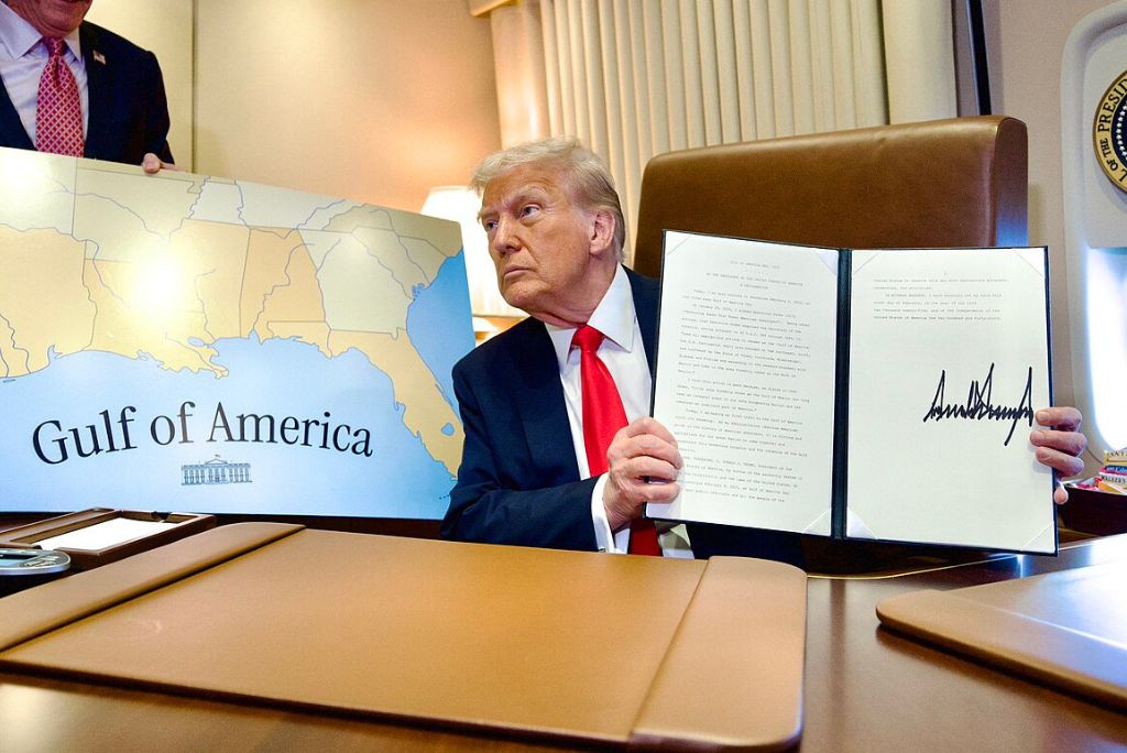 Donald Trump holds a signed document while presenting a map labeled Gulf of America during an official event in the Oval Office.