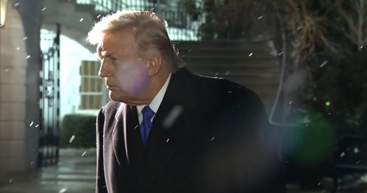 Donald Trump walks outside in the snow, wearing a dark coat and blue tie, with a serious expression on his face.
