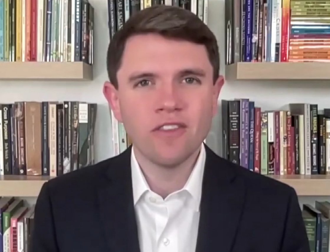 Man in a suit smiling in front of a bookshelf filled with various books, showcasing a professional and academic environment.