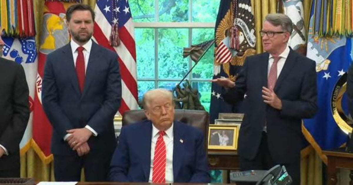Donald Trump seated in the Oval Office with advisors during a press event, surrounded by U.S. flags and presidential memorabilia.