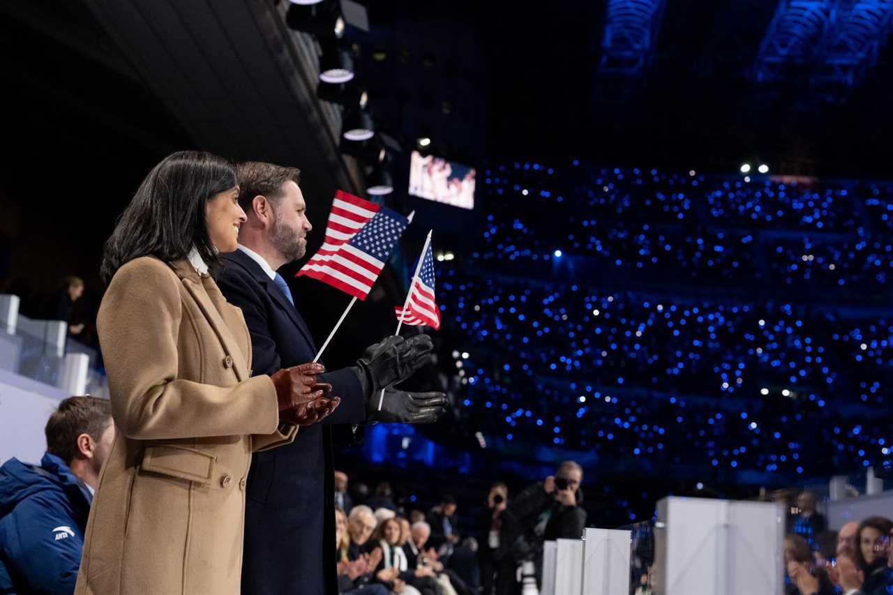 Two individuals hold small American flags while standing in front of a cheering crowd illuminated by blue lights during a significant event.