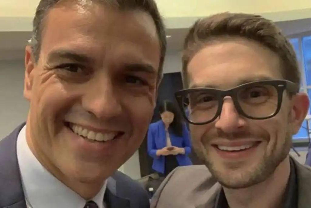 Two men smiling for a selfie, one in a suit and the other in glasses, with a blurred background of people in an indoor setting.
