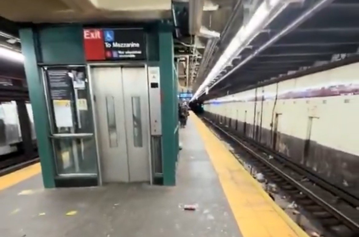 Subway station platform featuring an exit sign to the mezzanine, with empty tracks and minimal passenger activity.