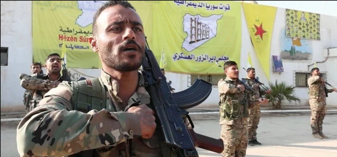 Members of a military group in camouflage uniforms perform a salute while holding rifles in front of colorful banners in a training area.