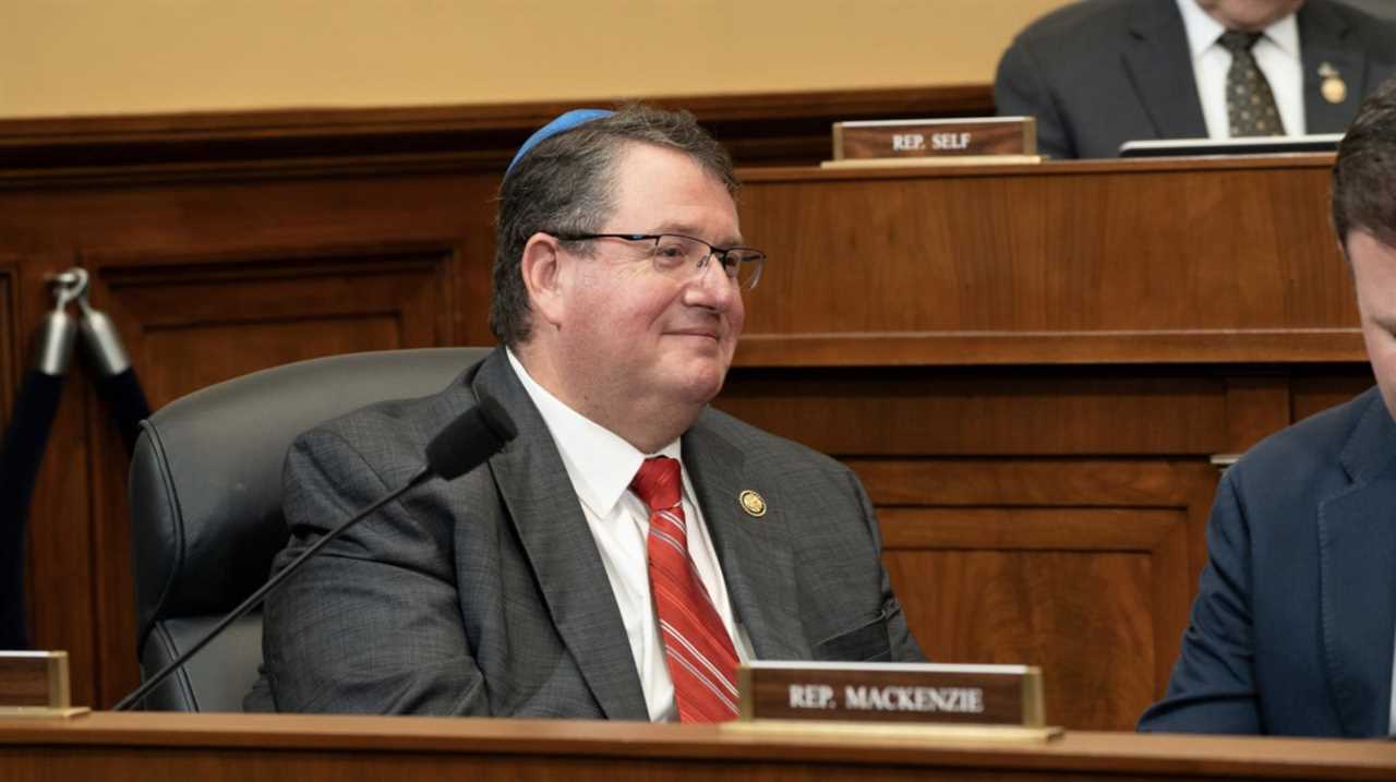 Representative Mackenzie smiles during a congressional meeting, wearing a suit and red tie, with fellow lawmakers in the background.