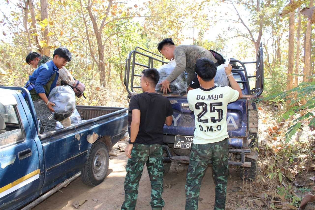 Group of individuals loading bags into the back of a blue pickup truck in a forested area during daylight.