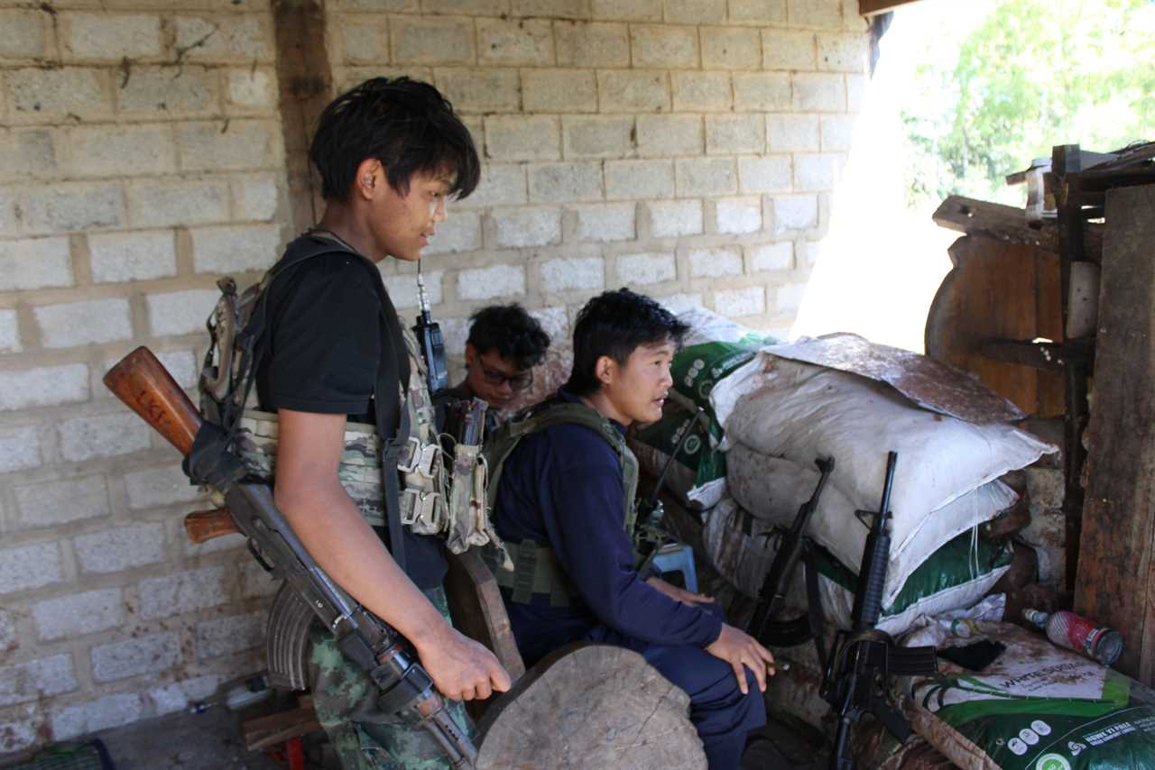 Young armed individuals are gathered inside a makeshift shelter, engaged in conversation amid military gear and supplies.