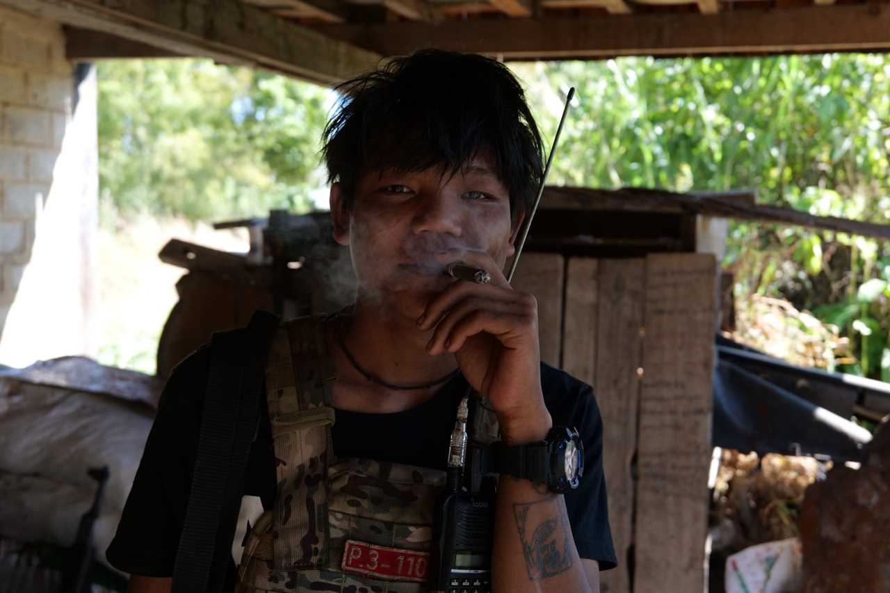 Young man in military attire smoking a cigarette, standing indoors with a radio, surrounded by rustic surroundings and greenery visible outside.