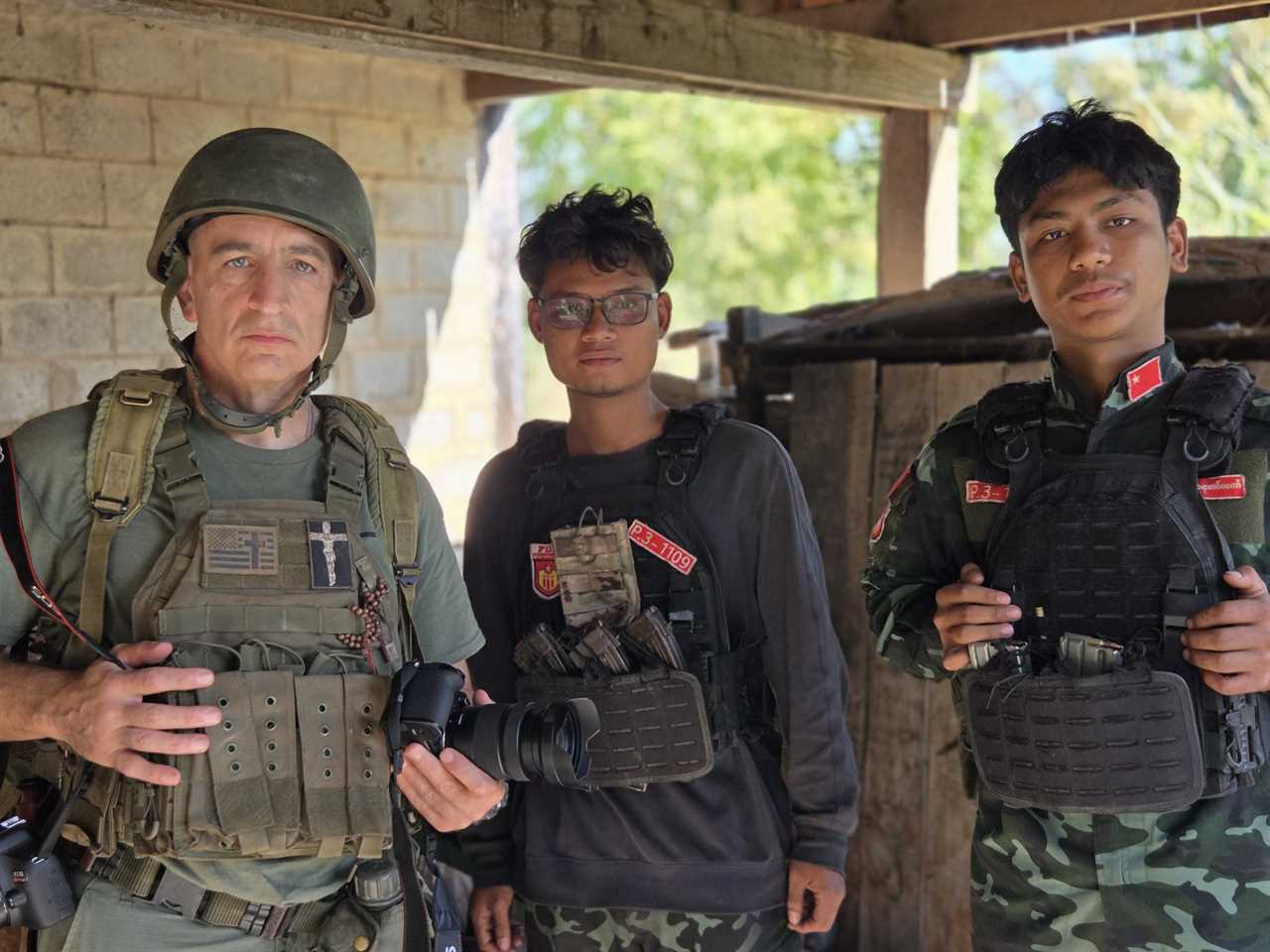 Three individuals in military attire pose together, with one holding a camera, inside a rustic shelter surrounded by greenery.