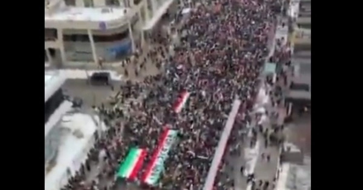 Aerial view of a large crowd participating in a protest, featuring flags and banners, demonstrating civic engagement and social activism in a city setting.