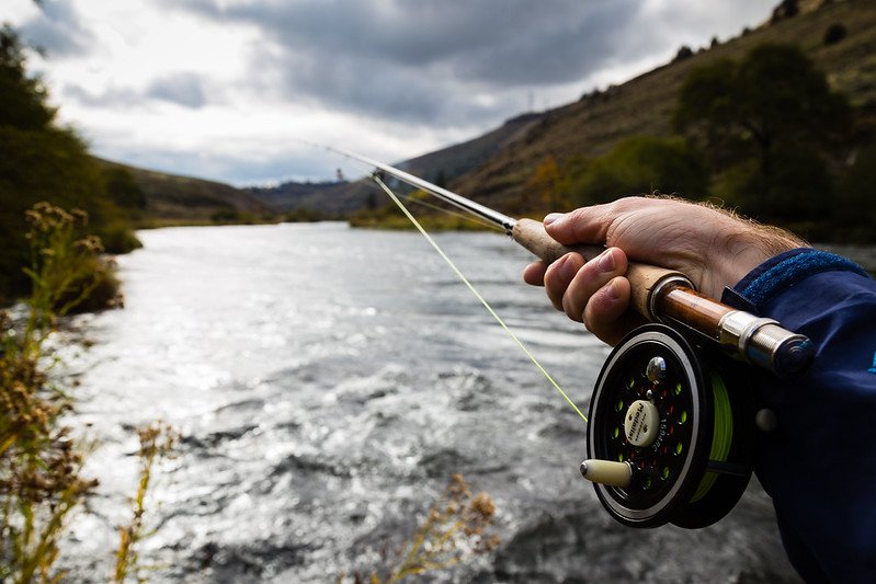 A person fly fishing on a river with a cloudy sky and rolling hills in the background.