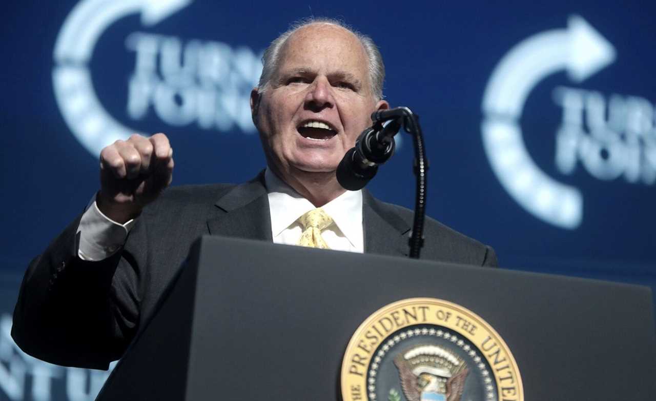 Rush Limbaugh speaking passionately at a Turnpoint USA event, gesturing emphatically while addressing the audience from a podium featuring the presidential seal.