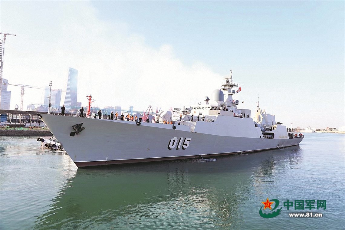 Modern naval vessel 015 docked at a port, showcasing military personnel and a bustling waterfront with construction in the background.