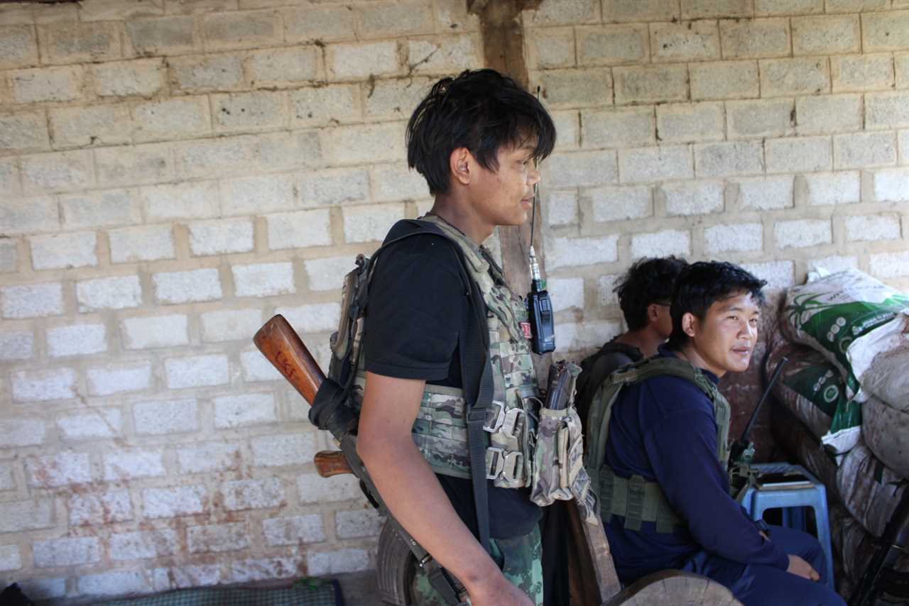 Two armed individuals in tactical gear inside a rustic shelter, one standing with a rifle and radio, while another sits nearby.