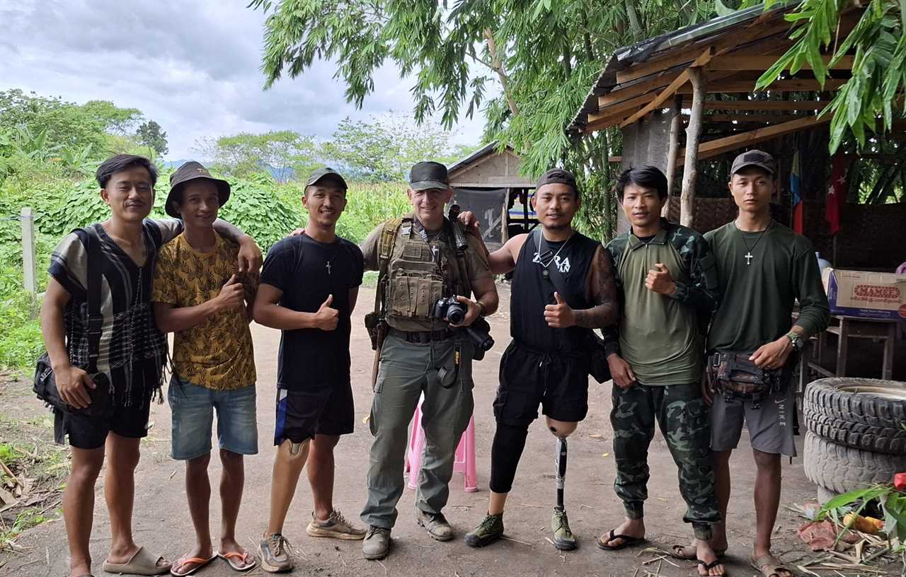 Group of seven individuals, including a soldier, posing outdoors in a rural setting with thumbs up, showcasing camaraderie and local culture.