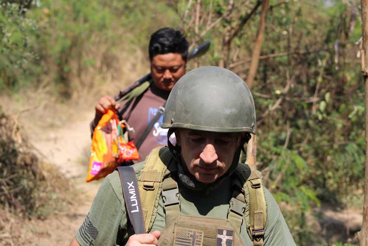 Soldier in military gear walks along a forest path, while a companion carries a bag, capturing a moment of outdoor exploration.