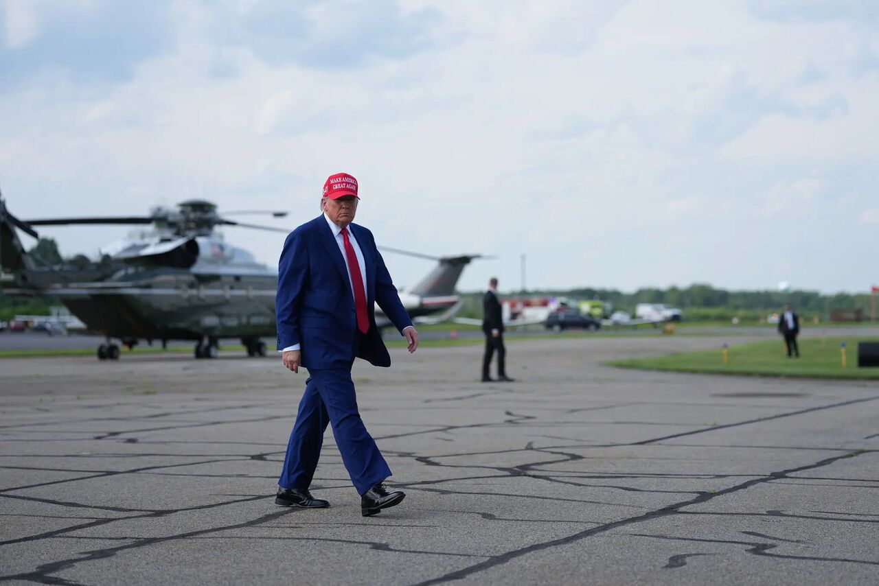Former President Donald Trump walks across the tarmac near helicopters, wearing a red cap and blue suit, amid a cloudy sky.