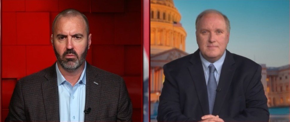 Two men in formal attire engaged in a serious discussion, with a backdrop featuring a prominent government building.
