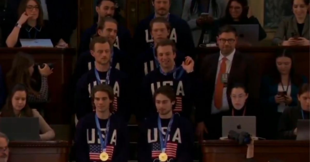 Athletes wearing USA sweatshirts and medals stand in a formal setting, surrounded by onlookers and media capturing the moment.