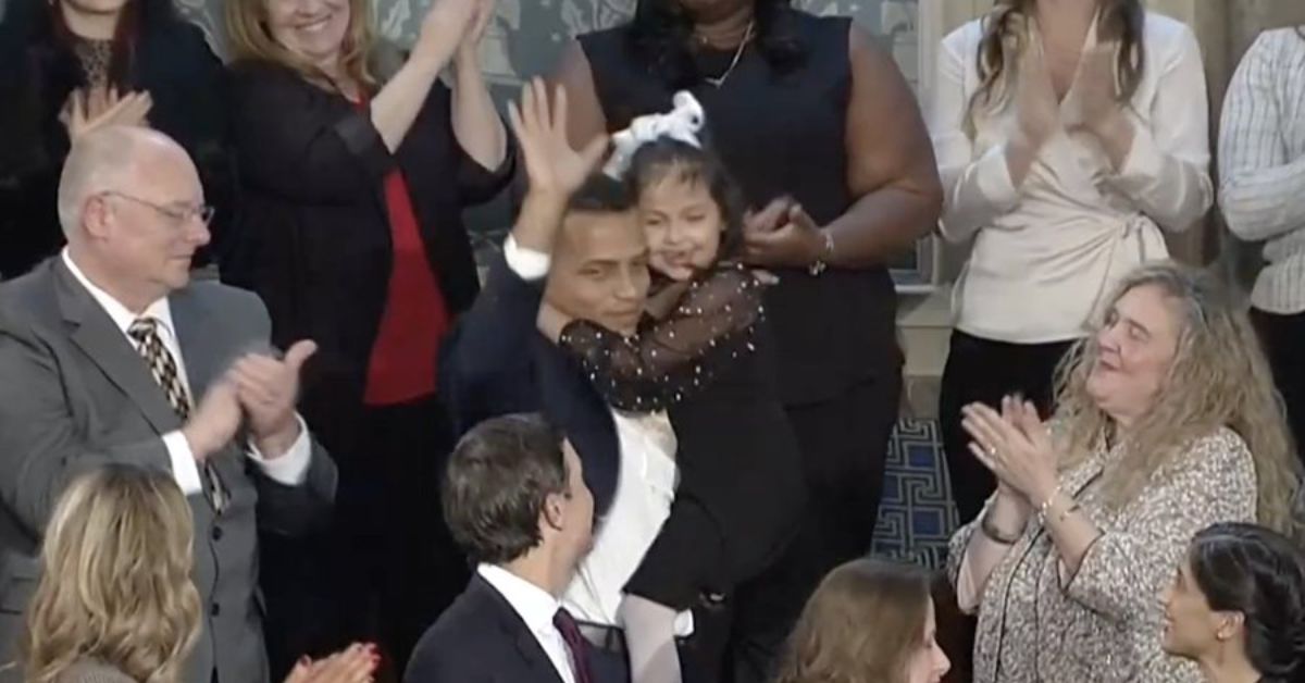 A smiling man holds a young girl while waving to an applauding audience at a public event, showcasing a moment of joy and celebration.