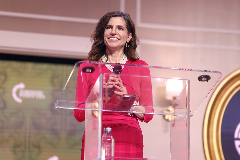 Woman in a red dress speaking at a podium with a microphone, smiling confidently during a presentation at a conference.