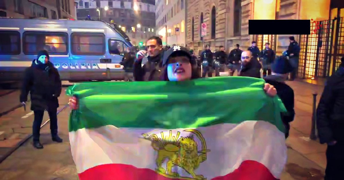 Protester holding a flag with a lion emblem during a demonstration, with police presence in the background and urban setting at night.