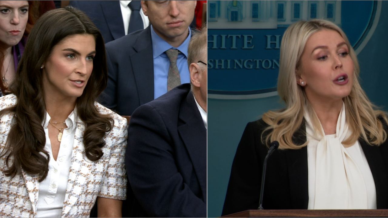 Two women in a press conference setting, one asking questions while the other speaks at the podium, highlighting media interaction in a political context.