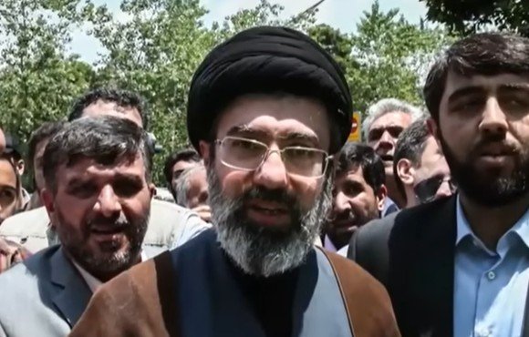 Religious leader addressing a crowd, surrounded by supporters, in a public outdoor setting with trees in the background.