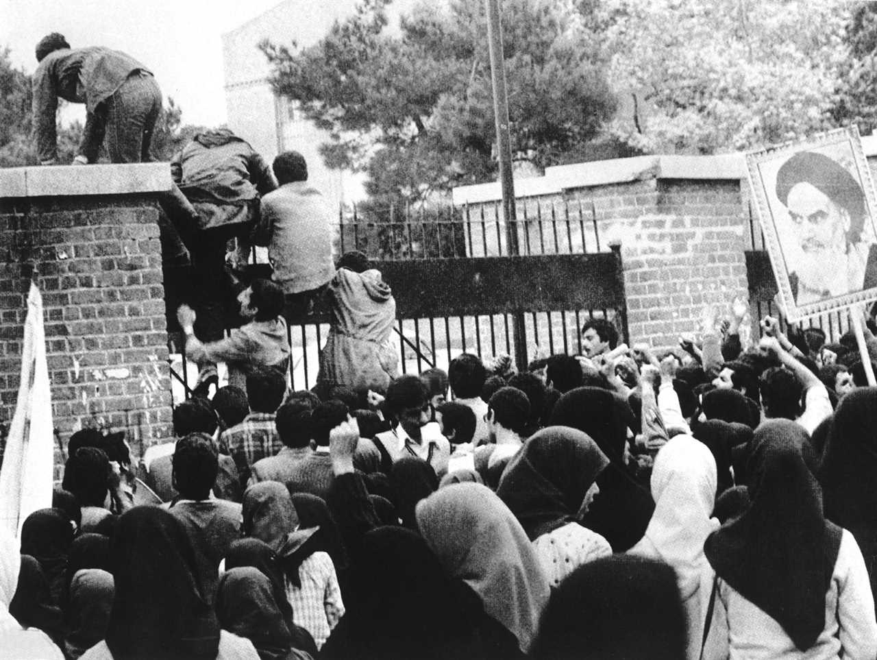 Crowd of protesters, some climbing a gate, during a historical demonstration with a portrait of a leader in the background.