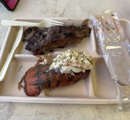 Grilled steak and lobster tail with garlic butter served on a tray, accompanied by a sugar packet and plastic utensils.