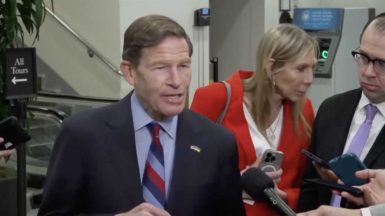 Senator Richard Blumenthal speaks to reporters while two individuals listen and take notes in a busy hallway.