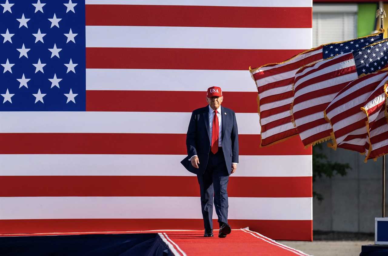 Man in a red USA hat walks on a red carpet in front of an American flag backdrop, flanked by waving flags.