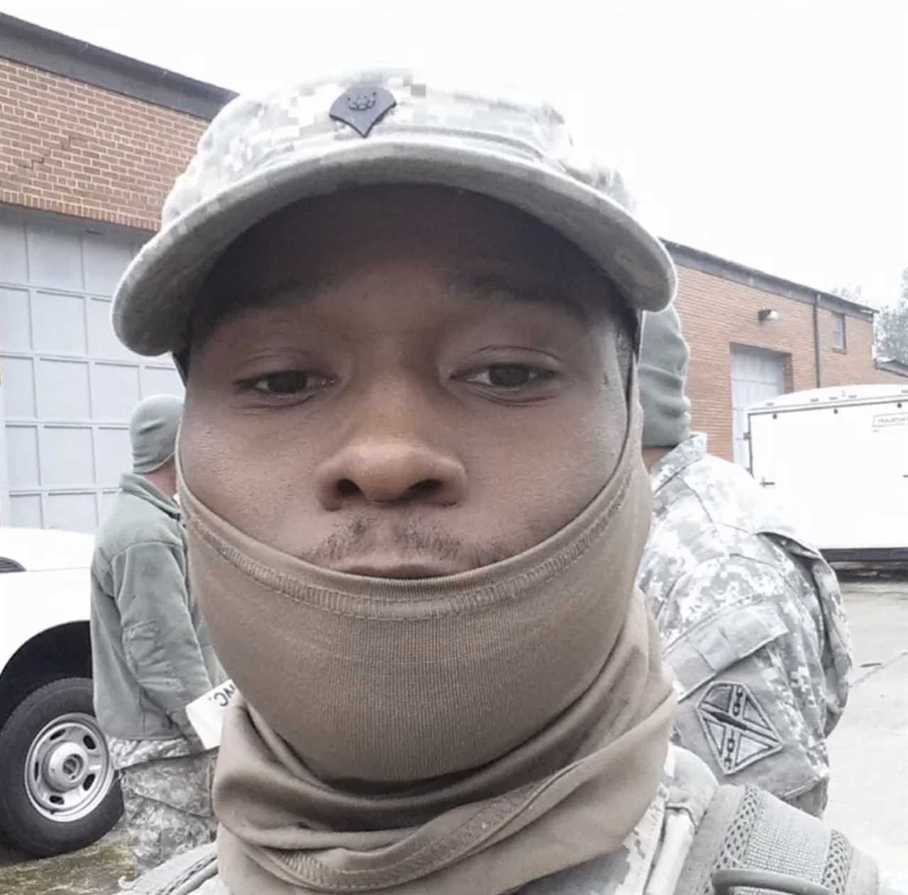 Soldier wearing military gear and a face mask poses for a selfie outdoors with fellow soldiers in the background.