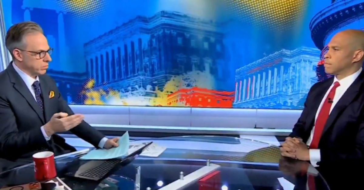 News interview featuring two men at a desk, discussing political topics, with a colorful backdrop of government buildings.