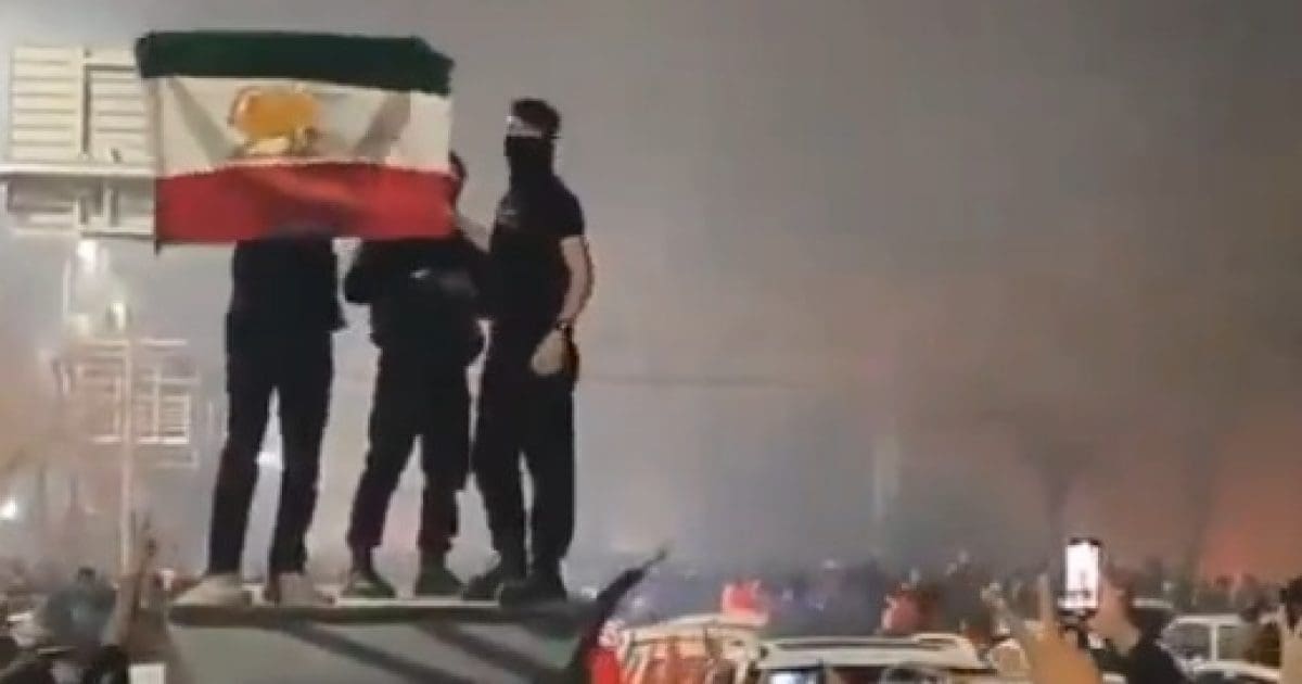 Protesters holding an Iranian flag stand atop a vehicle amidst a crowd during a demonstration, with smoke and excitement in the background.