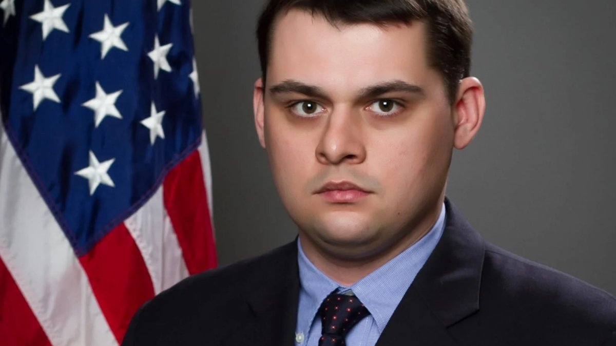 Portrait of a man in a suit with a serious expression, standing in front of an American flag backdrop.