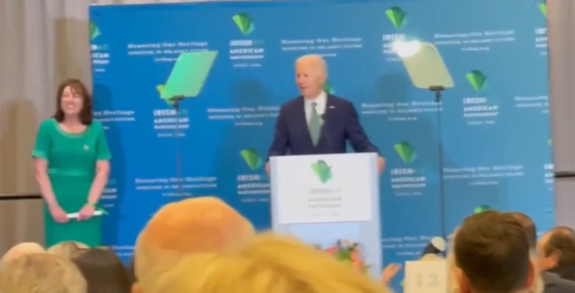 President Biden speaks at an event celebrating Irish American heritage, with a woman in a green dress standing beside him.