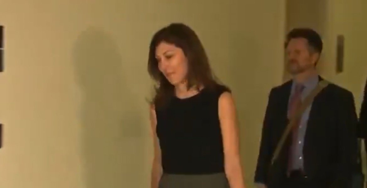A woman in a black top walks down a hallway, accompanied by a man in a suit, conveying a professional atmosphere.