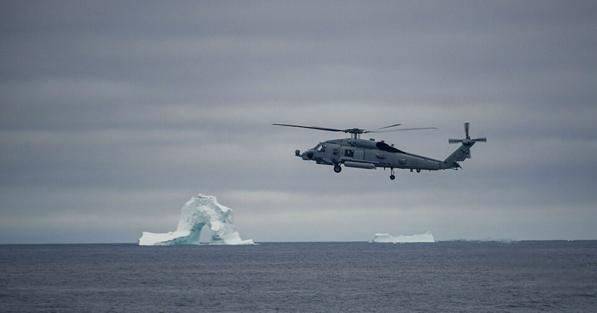 Military helicopter flying over ocean with icebergs in the distance under a cloudy sky.