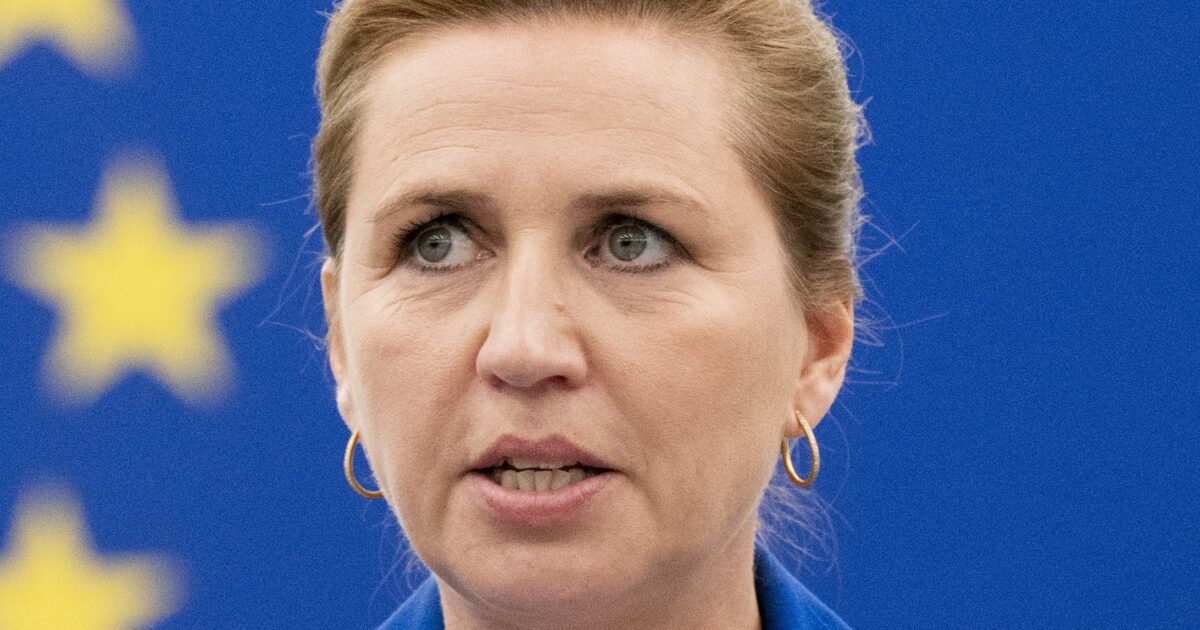 Close-up of a woman speaking at a podium with a blue background featuring European Union stars, showcasing her focused expression and professional attire.