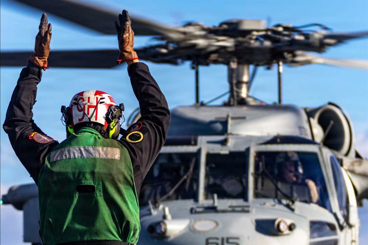A naval crew member signals a helicopter during landing operations on a ship, showcasing maritime aviation procedures.
