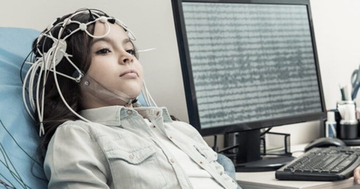 Child undergoing an EEG test with electrodes on her head, monitoring brain activity on a computer screen in a clinical setting.