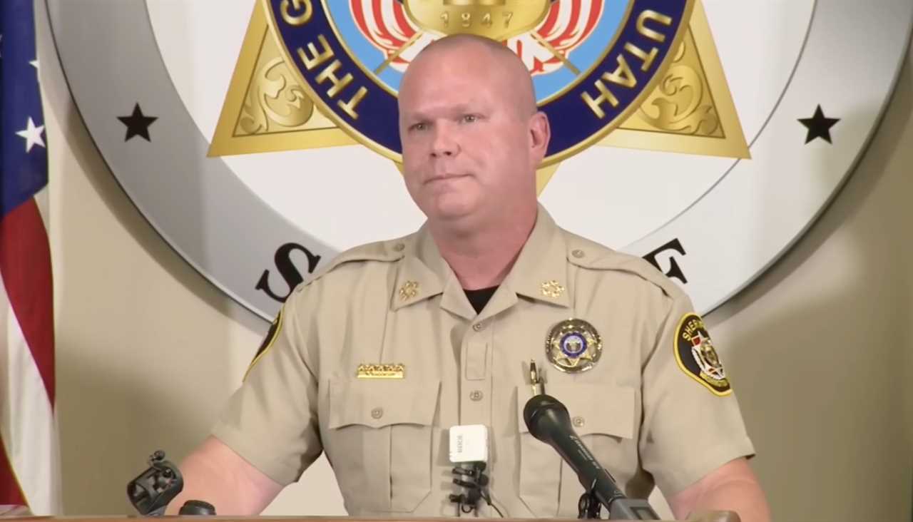 Sheriff addressing the media at a press conference, with a police badge visible and a law enforcement seal in the background.