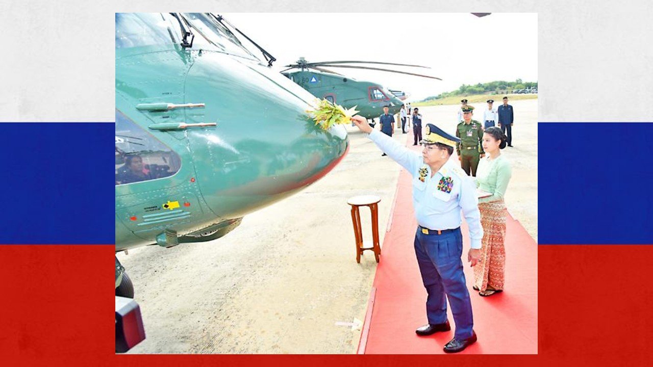 A military official performs a ceremonial blessing on a green helicopter during a formal event in front of attendees.