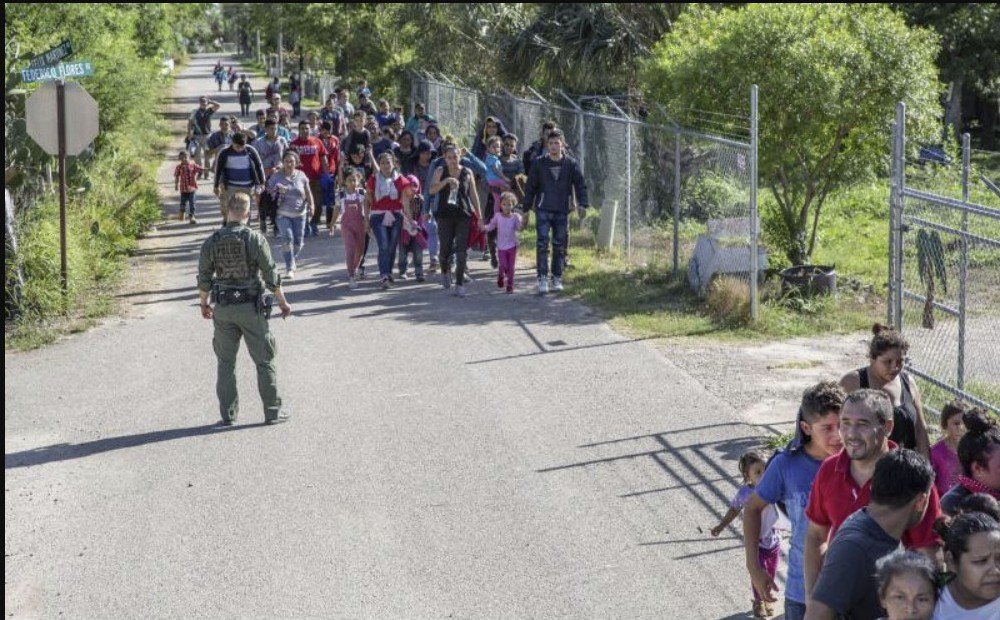 A group of migrants, including families and children, walks along a road while a border patrol agent stands nearby, highlighting immigration challenges.