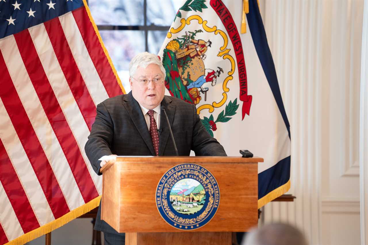 West Virginia official speaking at a podium with state flags in the background during a press event.